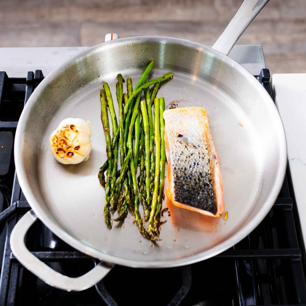 Stainless steel frying pan with seared salmon, asparagus and roasted garlic on a gas stove