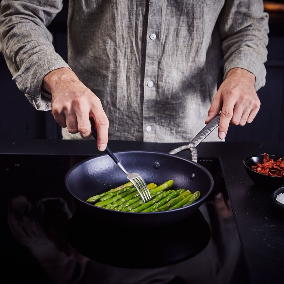 Man turning asparagus with a fork in a black frying pan on an induction cooktop