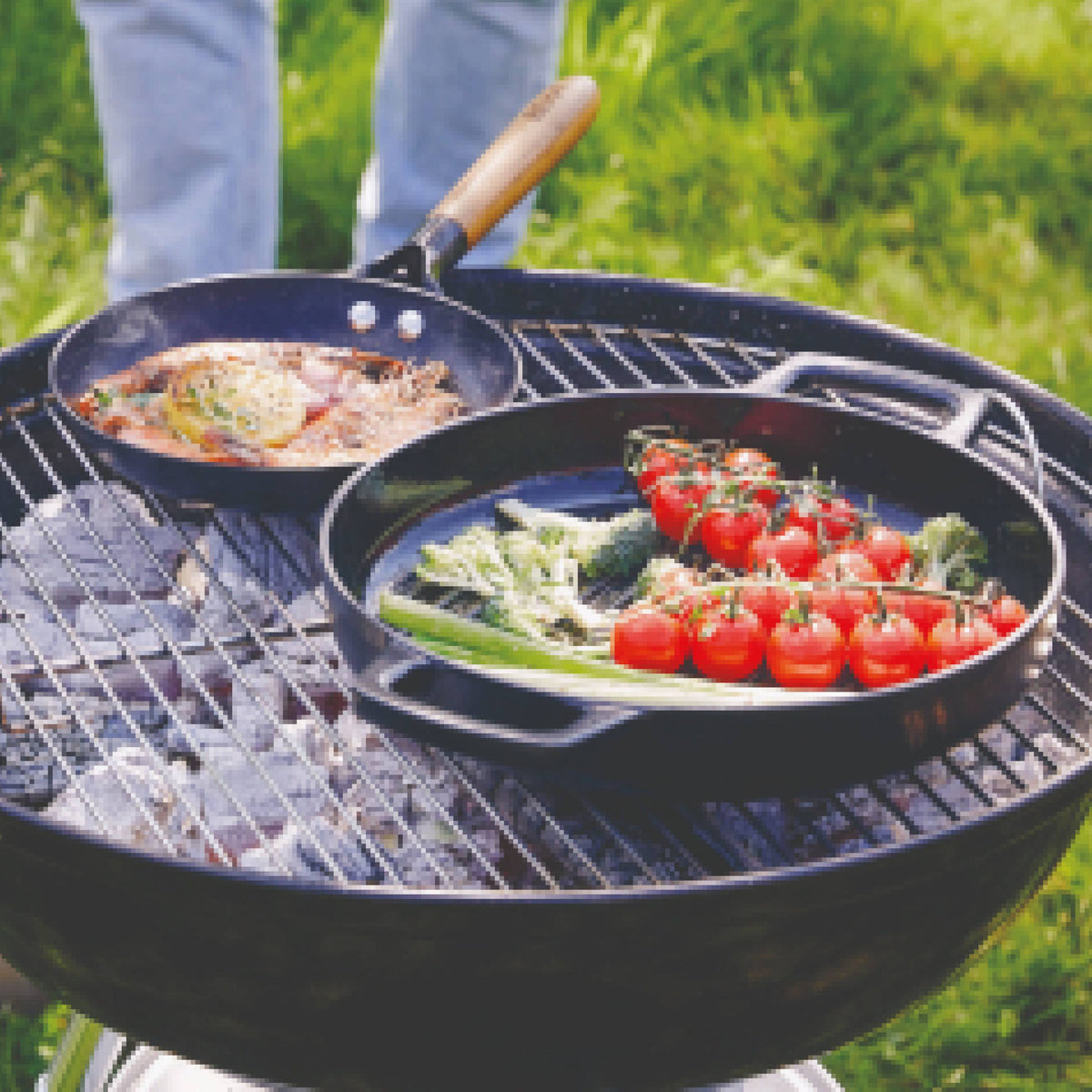 Black pans on an outdoor grill holding fish and grilled vegetables with cherry tomatoes