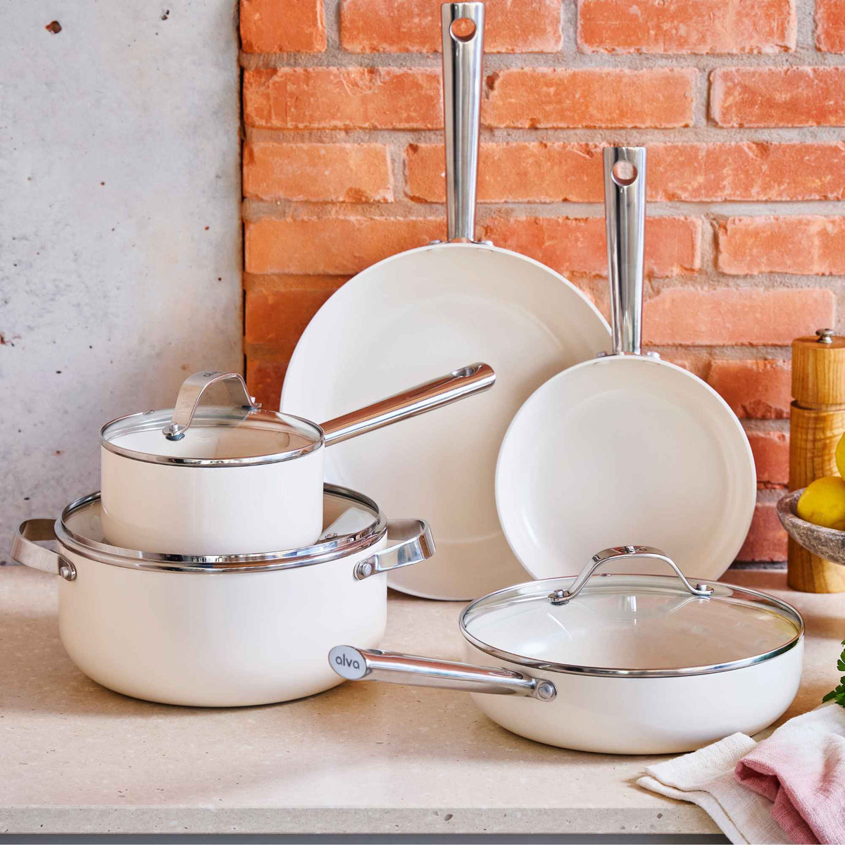 Alva cream ceramic nonstick cookware set with frying pans, a lidded saucepan and pots against a brick wall