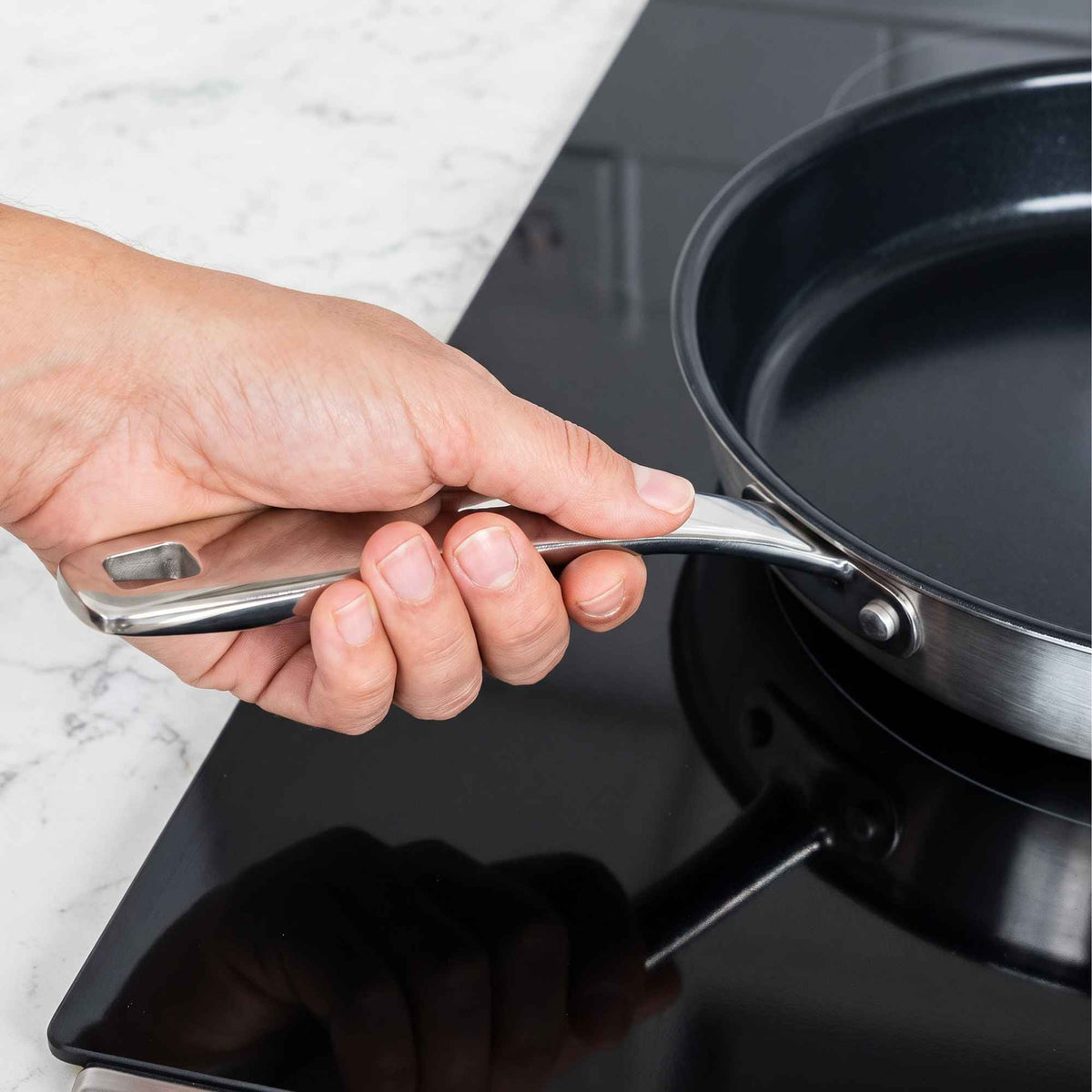 Hand gripping the stay-cool stainless steel handle of an Alva Maestro frying pan on a glass cooktop