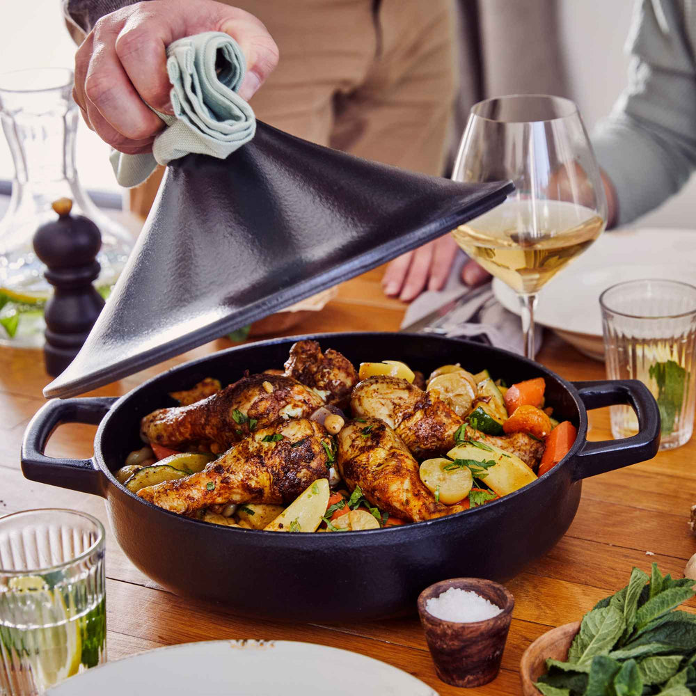 Chicken legs with potatoes and carrots in a black cast iron braiser, with the tagine-style lid being lifted off at the table