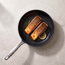 Overhead view of two salmon fillets with lemon and herbs in an Alva Maestro ceramic nonstick frying pan on a stone counter
