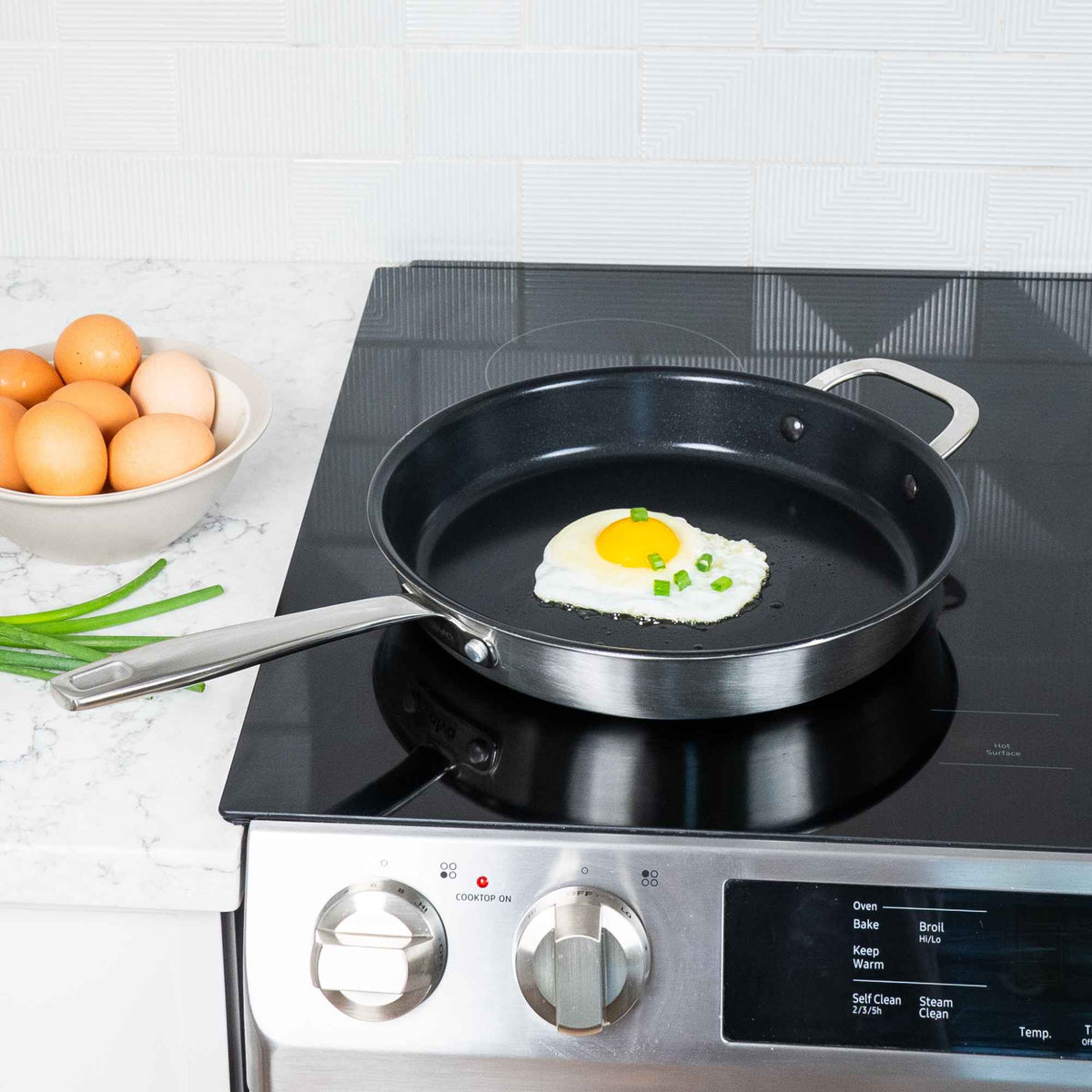 Fried egg with scallions in an Alva Maestro 5-ply ceramic nonstick frying pan on an induction range, eggs in a bowl beside it