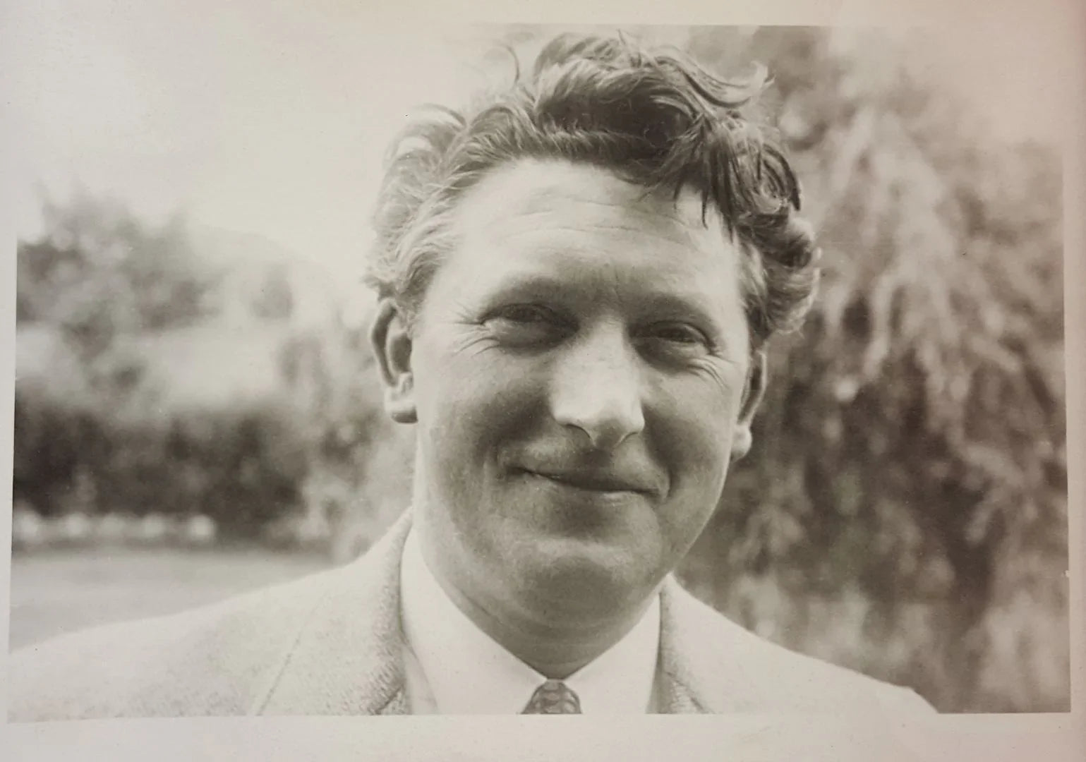 Vintage black and white portrait of a smiling man in a suit outdoors with blurred trees in background
