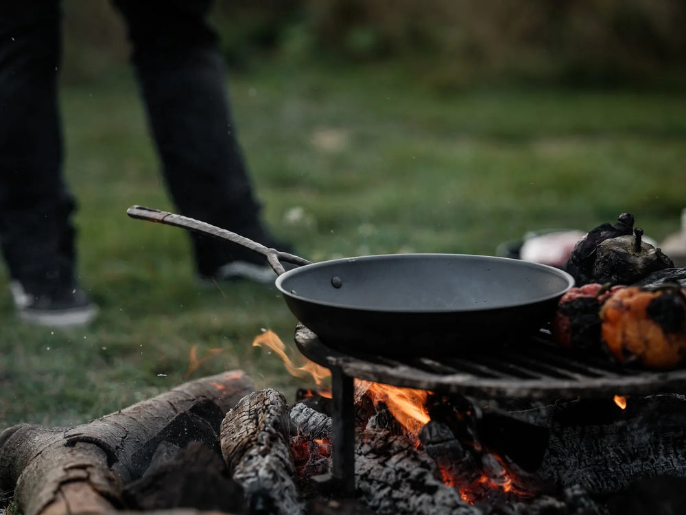 Black frying pan over open fire grill outdoors, with charred vegetables nearby.